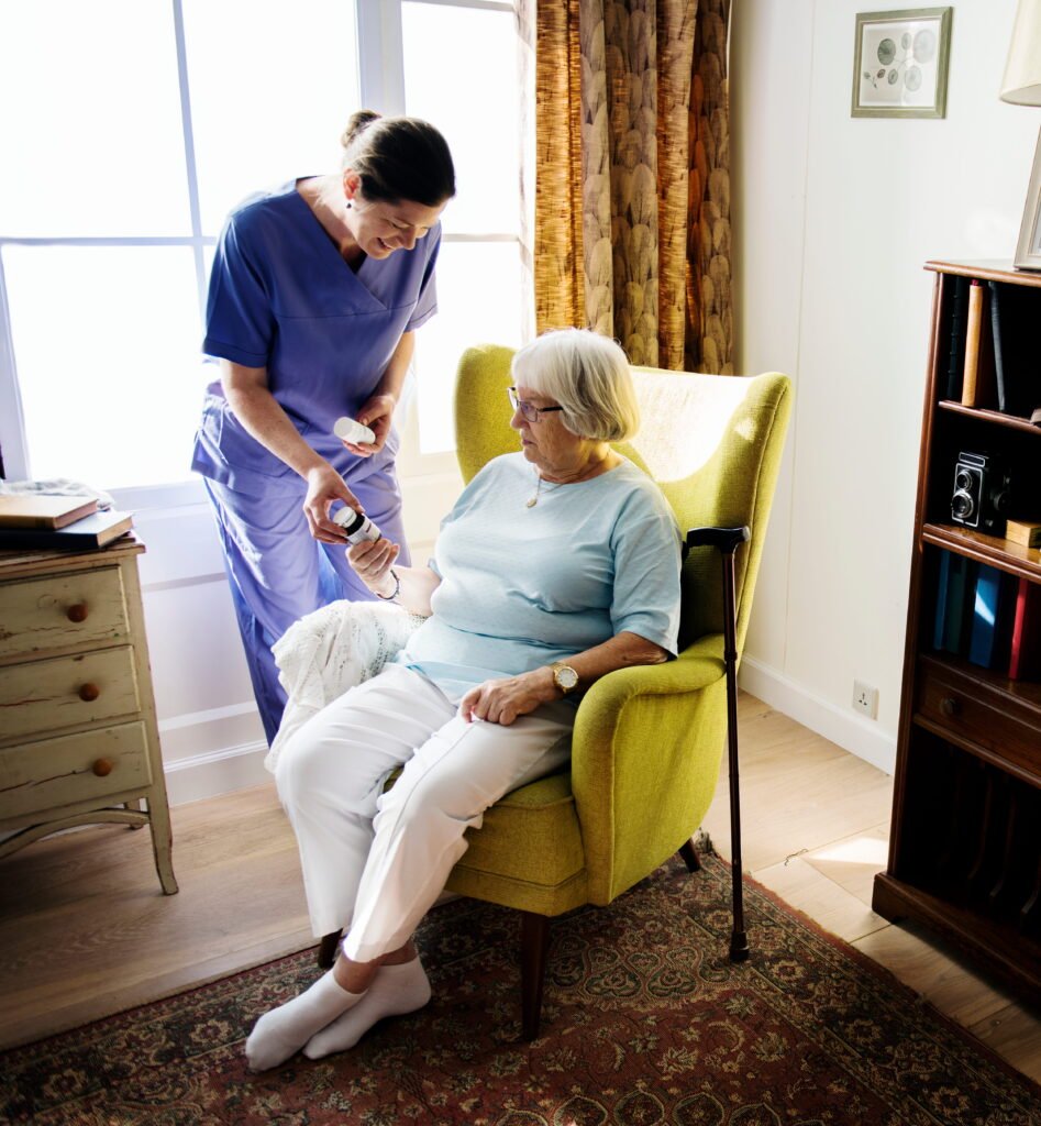 Nurse taking care of a senior woman in a living room. Elderly woman is sat on a yellow arm chair looking at a pot of pills the nurse is handing her